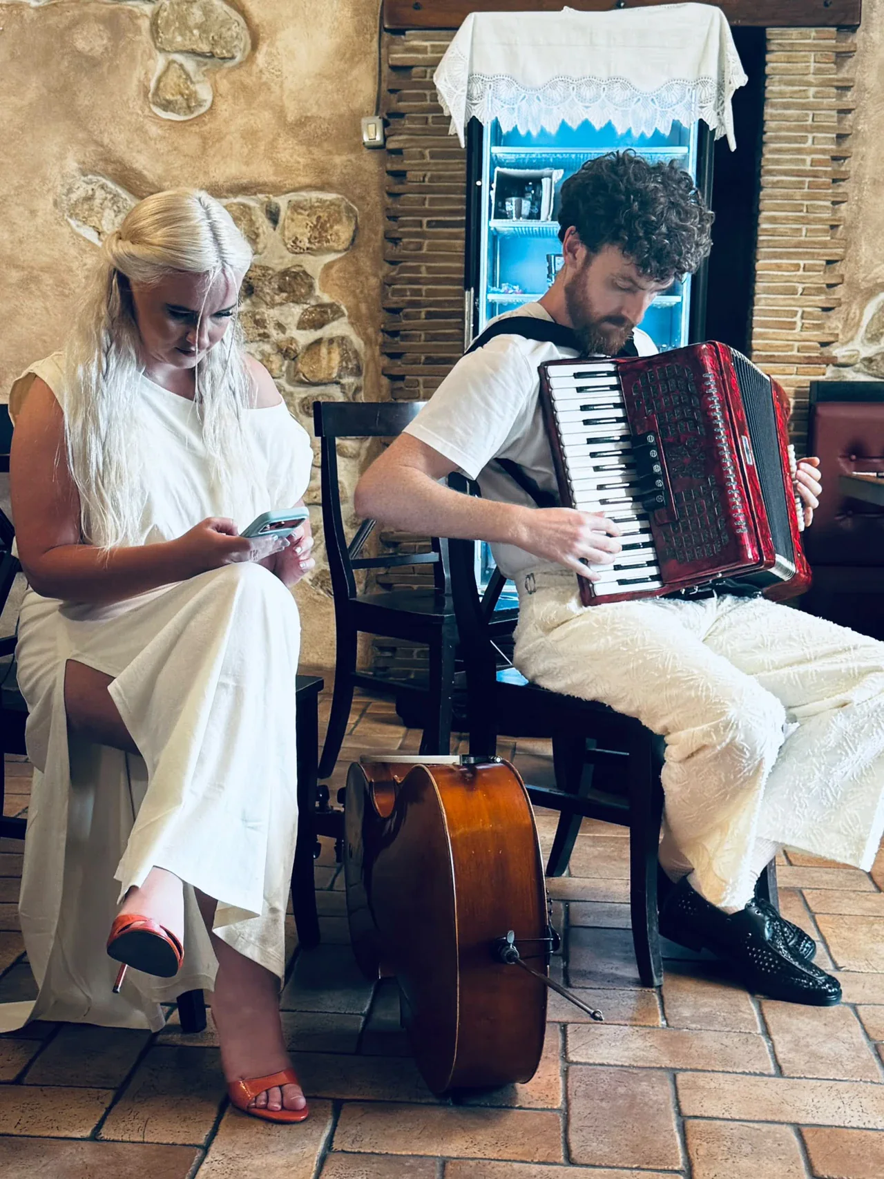 Due componenti dei Clean Bandit seduti in un interno: una persona consulta il telefono, l’altra suona una fisarmonica rossa; un violoncello è appoggiato davanti alle sedie.